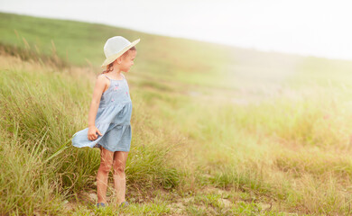 Naklejka na ściany i meble a little girl in a hat is standing in the middle of the field and looking at the sunlight, and the wind is blowing her dress