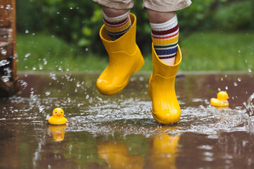 A small child in rainbow-colored socks and yellow rubber boots jumps through puddles and plays with...