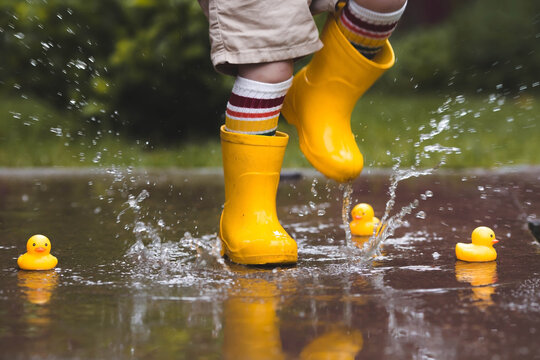 A small child in rainbow-colored socks and yellow rubber boots jumps through puddles and plays with yellow rubber ducks. Legs close-up. A picture of summer and autumn holidays. A child in the rain.