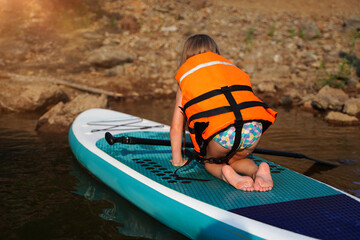 European child girl in a life jacket sits on a SUP board floating on the lake