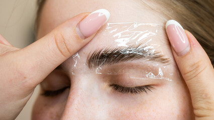 The master uses a plastic film during lamination of the eyebrows. - obrazy, fototapety, plakaty