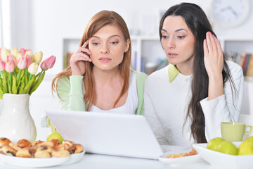 Portrait of two young women looking at laptop while sitting at kitchen table with cookies