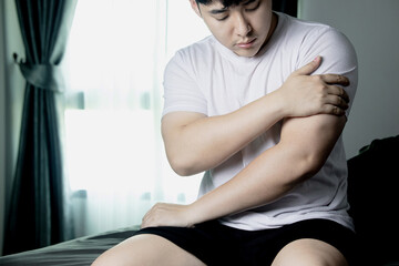 Shoulder pain, arm pain, man with musculoskeletal problems, concept of health care and medicine Asian man sitting in bed and holding a painful arm muscle with one hand. from sleeping the wrong way 