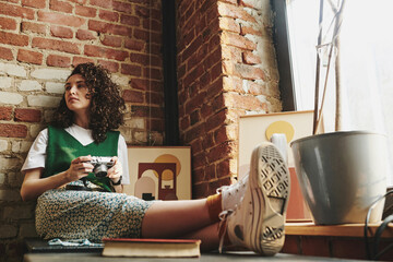 Young woman in casualwear holding retro photocamera while sitting against red brick wall by window...