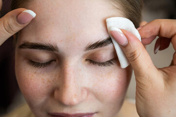 The master processes the eyebrows of a Caucasian woman with a cotton pad before laminating the...