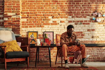 Young relaxed black man in casualwear looking through contacts or playlist in his smartphone while...
