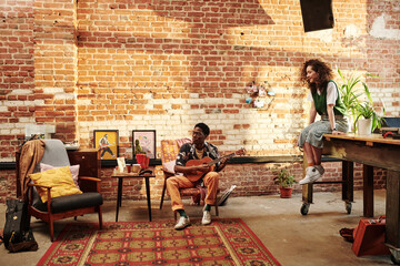Young female in casualwear sitting on table and listening to her African American boyfriend playing...