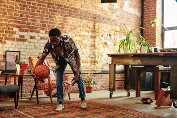 Young African American man in casualwear playing with ball on carpet in spacious living room of...