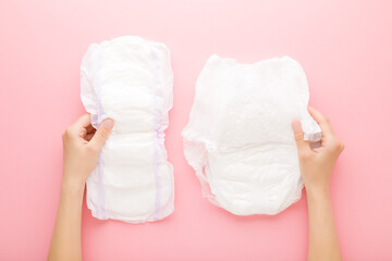 Young adult woman hands holding white big size sanitary towel and diaper pant on pastel pink background. Closeup. Hygiene product for urinary incontinence or after childbirth. Top down view. - obrazy, fototapety, plakaty