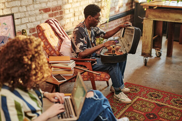 Young African American man opening case with violin while sitting in armchair in front of his girlfriend with laptop on her knees