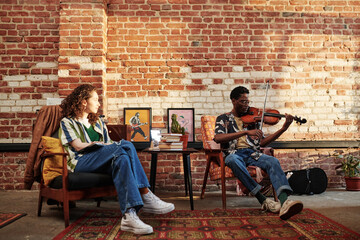 Young woman looking at her African American boyfriend playing violin while sitting in armchair in living room of loft apartment - obrazy, fototapety, plakaty