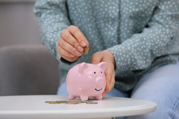 Young woman putting coin into piggy bank at table indoors, closeup