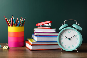 Alarm clock and different stationery on wooden table near green chalkboard. School time