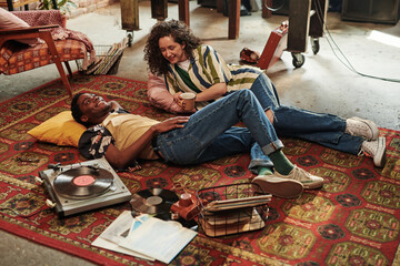 Happy young couple in stylish casualwear listening to vynil disks and talking while relaxing on red carpet on the floor of living room - Powered by Adobe