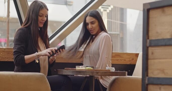 Two female friends posing for selfie in restaurant before eating meal, then they choose the best photo. Caucasian and Moroccan woman taking a selfie in a coffee with mobile phone.