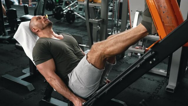 Concentrated muscular middle-aged male bodybuilder wearing a tight t-shirt and shorts exercising his leg muscles on a leg press. High quality 4k footage