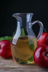Jug of tasty juice and fresh red apples on wooden table against black background