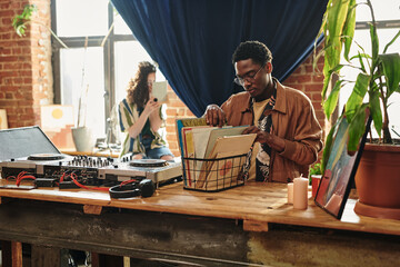 Young African American man looking through his collection of vynil disks in box standing on table...