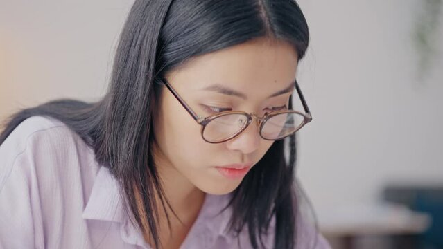 Asian woman in glasses studying at home, writing in notebook, distance education