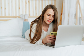 Freelance woman texting over cellphone and having a video call chat online shopping, lying on the white bed. Happy relaxed girl woking from home office. Distance learning online education and work.