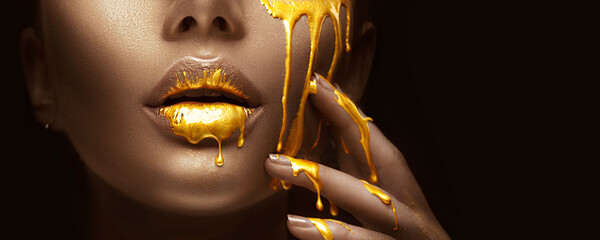 Gold Paint smudges drips from the face lips and nails, lipgloss dripping from sexy lips, golden liquid drops on beautiful model girl's mouth, gold metallic skin make-up. Beauty woman makeup close up.