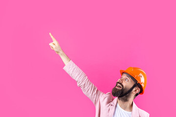 Portrait of young construction engineer wear orange hard hat, in a pink jacket standing on red studio background. The man points with his hand.