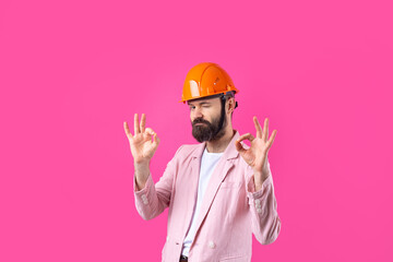 Portrait of young construction engineer wear orange hard hat, in a pink jacket standing on red studio background. The man points with his hand.