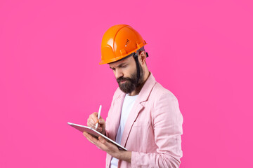 Portrait of young construction engineer wear orange hard hat, in a pink jacket standing on red studio background. A man with a tablet in his hands.