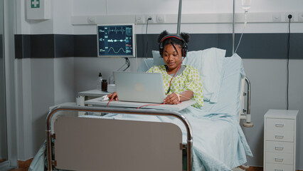 Patient with sickness using headphones to listen to music while sitting in hospital ward bed. Young woman looking at laptop and listening to audio sounds on headset. Adult with disease