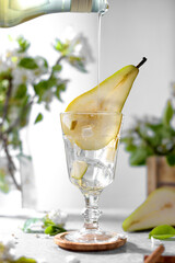 Pear syrop is pouring in a glass with pear cocktail decorated with fresh fruit and flowers on light gray background. Summer cold pear drink lemonade with ice on the table, Mocktail with flowering twig
