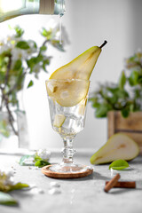 Pear syrop is pouring in a glass with pear cocktail decorated with fresh fruit and flowers on light gray background. Summer cold pear drink lemonade with ice on the table, Mocktail with flowering twig
