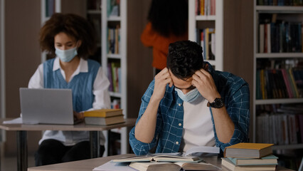Tired overworked young arab male student in protective mask sits at desk in library does homework...