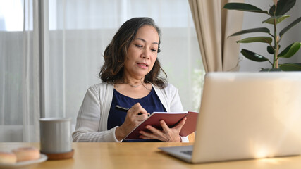 Concentrated middle aged businesswoman watching online webinar on laptop computer and making notes on notebook while remote working from home