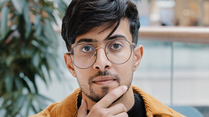 Headshot young pensive thoughtful indian man holding hand on chin thinking deep in thought in...