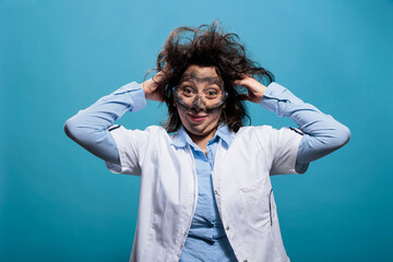 Crazy chemist with goofy appearance feeling guilty for explosion resulted from failed chemical experiment. Mad scientist with messy hair, dirty face and dumb expression standing on blue background. - obrazy, fototapety, plakaty