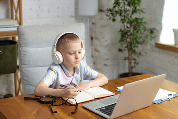 Naklejka premium One cute little boy, school age kid listening online lesson, using laptop at home interior. Education, childhood, people, homework and school concept.
