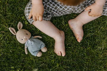 Closeup of baby sitting on grass with barefoot feet next to a  crocheted bunny toy.
