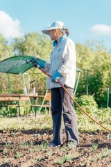 A woman in a hat waters the beds with a sprayer connected to a hose.