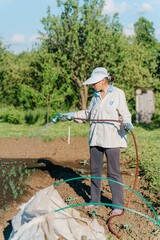 A woman in a hat waters the beds with a sprayer connected to a hose.