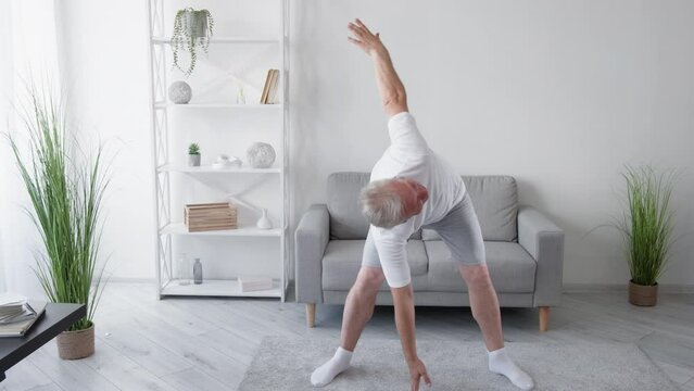 Morning fitness. Home pilates. Yoga training. Inspired smiling athletic middle-aged man performing twists and turns stretching back muscles exercise in light living room interior.