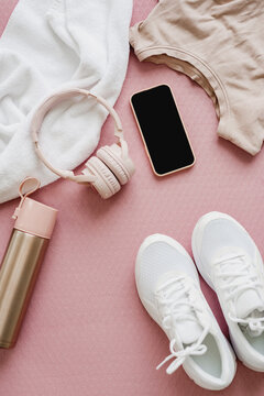 Flat lay of female sports clothing, smartphone, headphones, and a bottle of water on pink background.