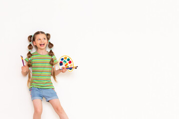 Drawing for schoolchildren. A little girl in a top and shorts with a palette of paints and brushes shows your advertisement on a white isolated background. Copy space.