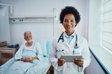 Happy black doctor uses touchpad while visiting her patient at hospital ward and looking at camera.