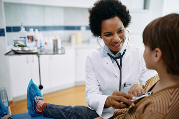 Happy black female pediatrician examining little boy with stethoscope during medical appointment at clinic.