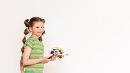 Drawing for schoolchildren. A little girl in a top with a palette of paints and brushes shows your advertisement on a white isolated background. Copy space.