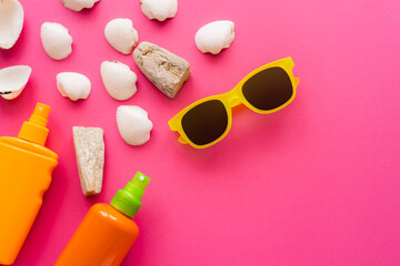 Top view of sunglasses near sunscreens and seashells on pink background.