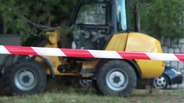 Close-up of the fence with a red and white ribbon against the background of yellow construction equipment which means that construction work is dangerously carried out there