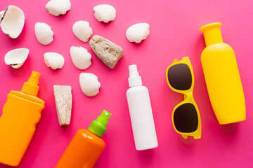Top view of sunglasses and sunscreens near white seashells on pink background.