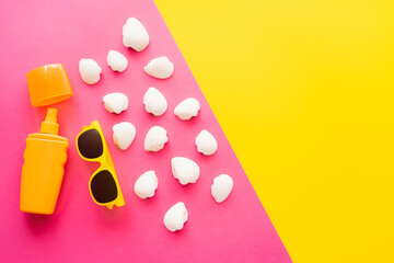 Top view of sunblock near sunglasses and seashells on pink and yellow background.