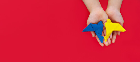 two doves made of origami paper in yellow and blue of colors of Ukrainian flag in children's hands on red background
